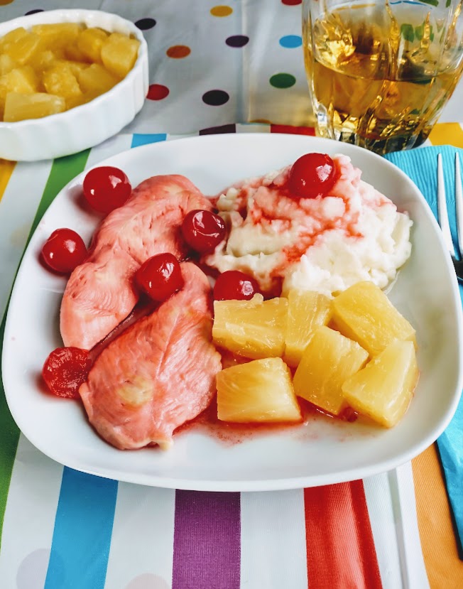 a square white plate with cherry chicken tenders, whole cherries mashed potato and pineapple and a glass of apple juice