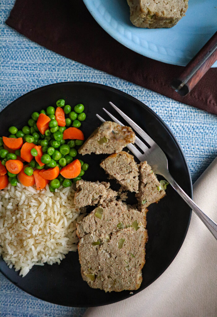 a slice of ground turkey meat cut up on a dark plate served with carrots peas and rice